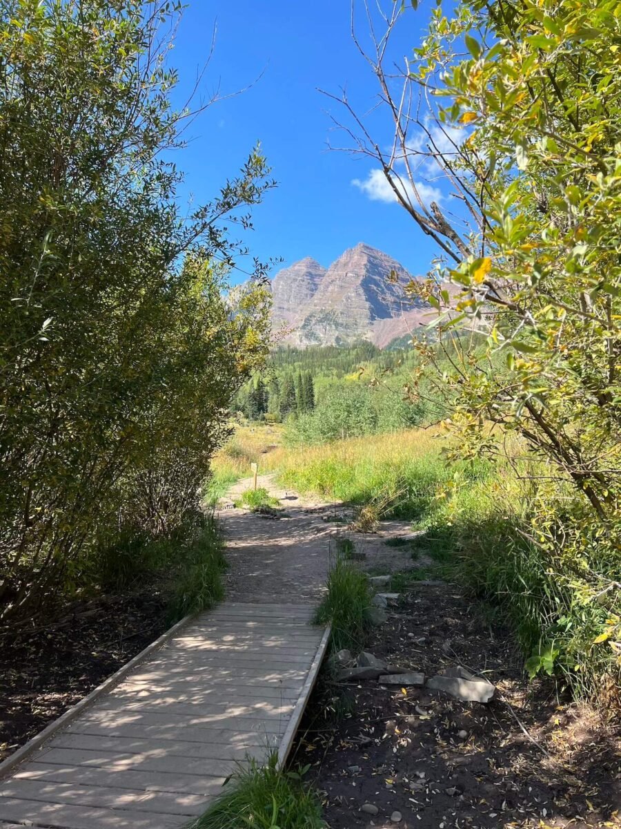 Pathway framed by lush greenery leading to the iconic Maroon Bells peaks under a bright blue sky. A scenic and popular hiking destination near Denver, Colorado.
