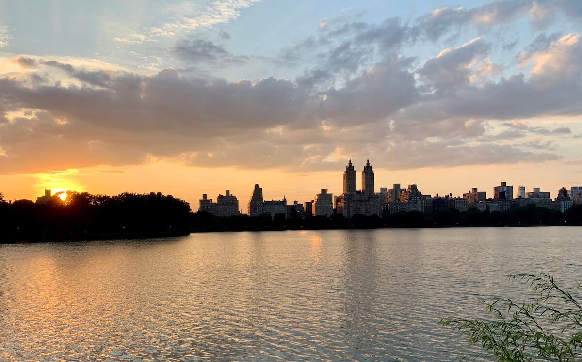 The Jacqueline Kennedy Onassis Reservoir in Central Park at sunset, with the evening sky casting a warm glow over the water and the silhouette of the Manhattan skyline in the background.