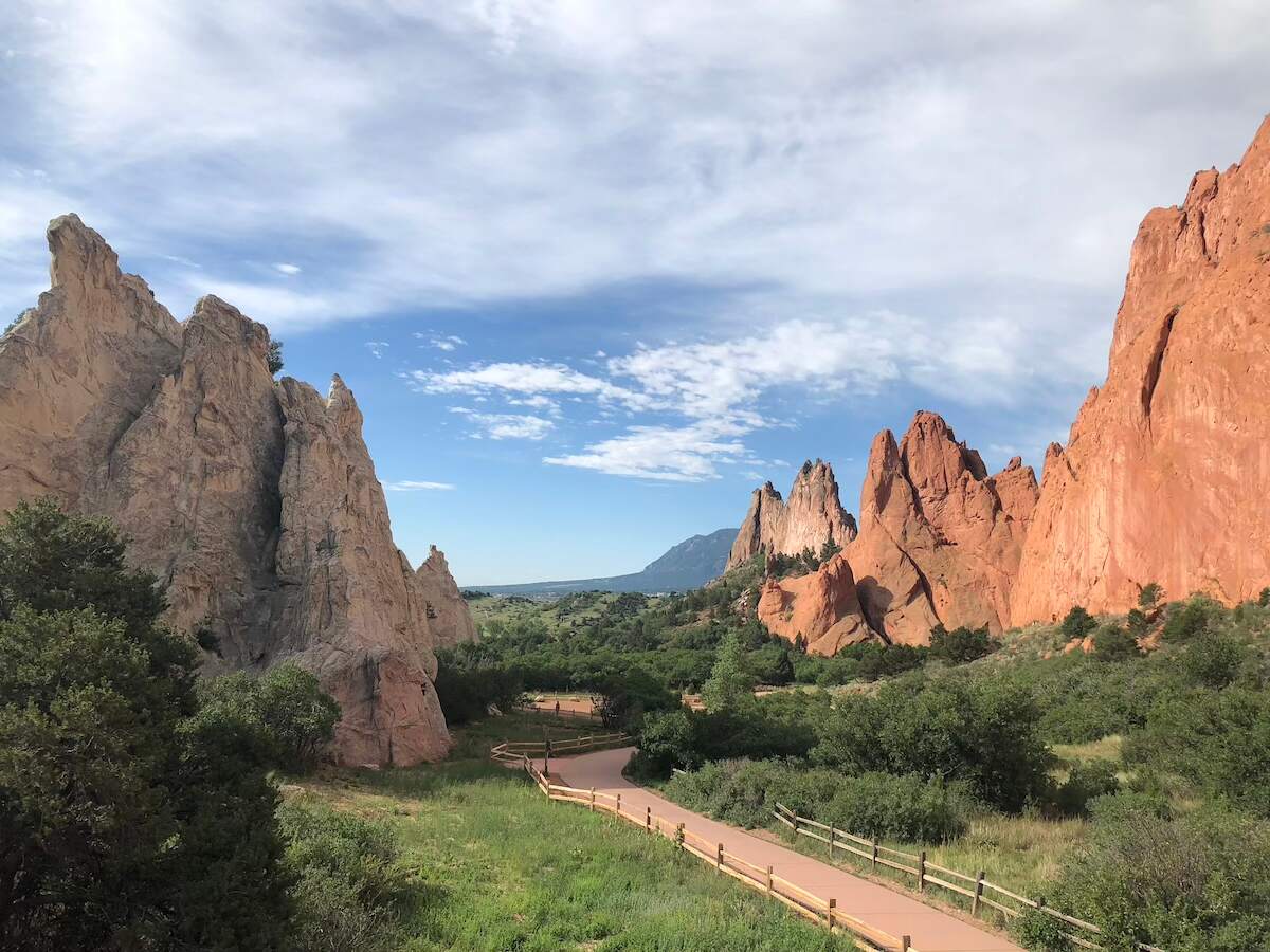 Pathway winding through the towering red rock formations of Garden of the Gods in Colorado Springs, with lush greenery and a blue sky with scattered clouds. A popular road trip destination from Denver, Colorado.