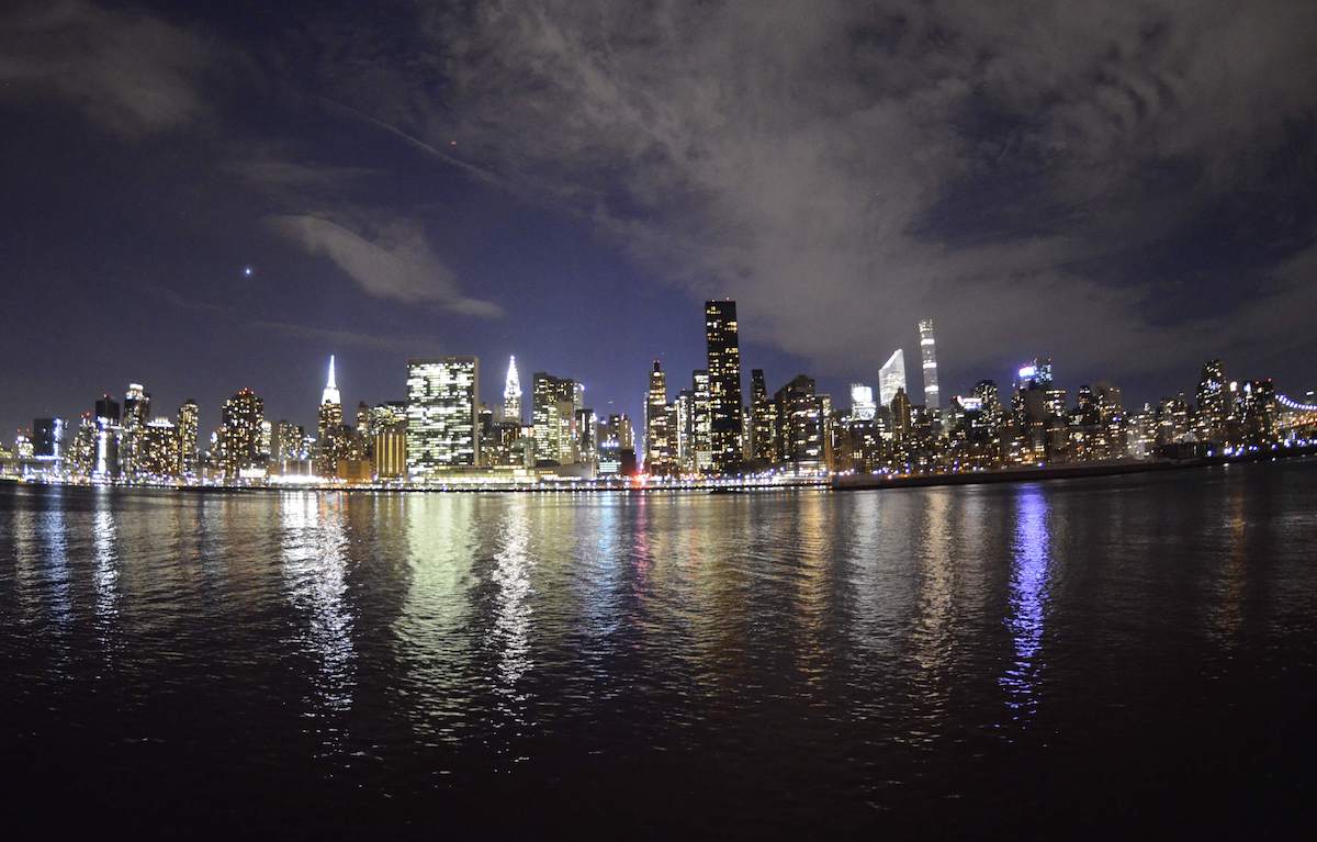 The Manhattan skyline viewed from Gantry Plaza State Park in Queens, NYC at night, with city lights reflecting on the East River under a partly cloudy sky.