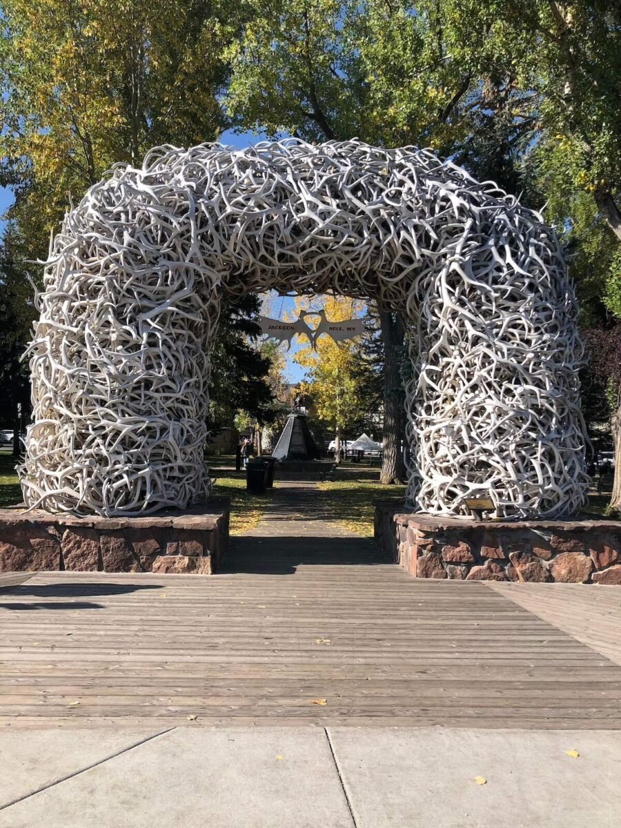 Arch made of intertwined antlers with a sign reading 'Jackson Hole, WY' in the center, located in a park with trees and a statue in the background. A unique and iconic road trip destination from Denver, Colorado.