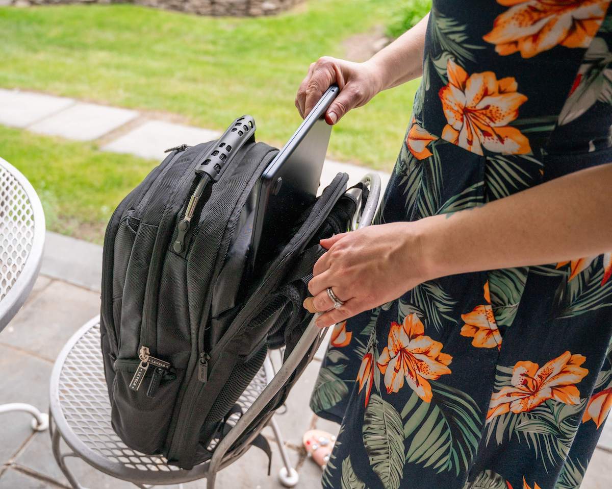 A person placing a laptop into the LEVEL8 Atlas Laptop Backpack. The person is wearing a dress with a dark background and orange floral print, and a silver ring on their left hand. The scene is set outdoors with a grassy background and a white metal patio table and chair.