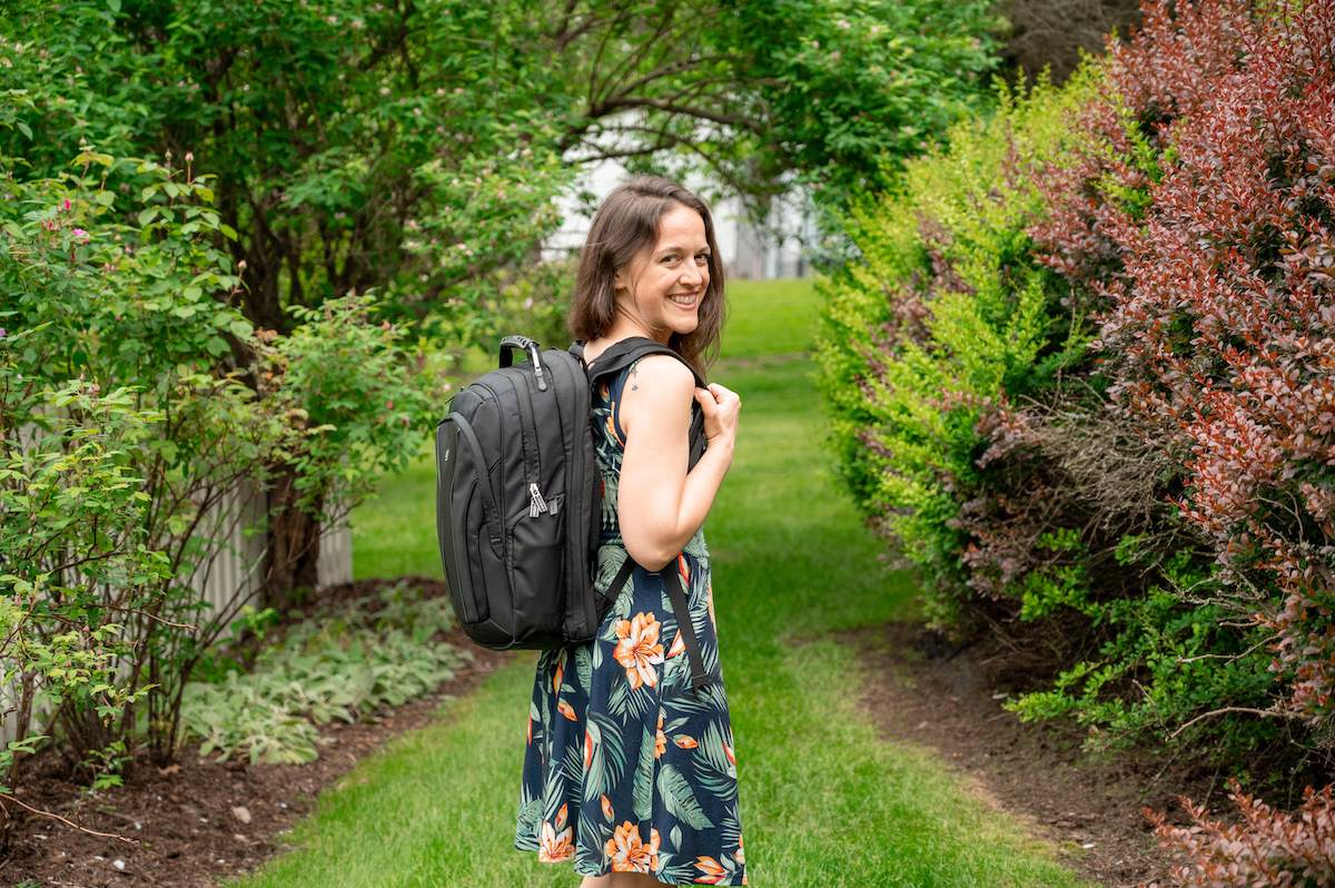 woman in a blue dress smiling at the camera while holding onto the straps of the LEVEL8 Atlas Laptop Backpack