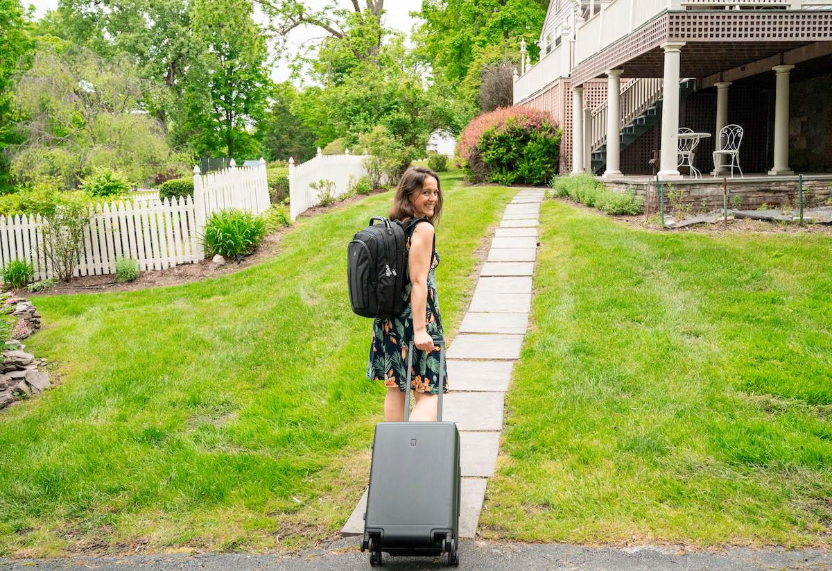 A woman with a LEVEL8 Atlas Laptop Backpack on her back and pulling a LEVEL8 Luminous Textured carry-on suitcase walks along a stone path in a grassy yard. She is smiling and looks back towards the camera, showcasing her travel essentials. The background features a white picket fence and a house with a porch and outdoor furniture, highlighting a scene perfect for travel with cabin baggage and an aluminum suitcase from the LEVEL8 luggage set.