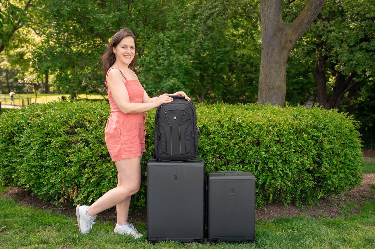 A woman in a coral romper stands outdoors on grass beside a row of three black LEVEL8 luggage pieces: an Atlas Laptop Backpack on top of a medium-sized Luminous Textured suitcase, with a larger suitcase to the left and a smaller carry on suitcase to the right. She is smiling and has her hand resting on the backpack. There is greenery and trees in the background behind the luggage set.
