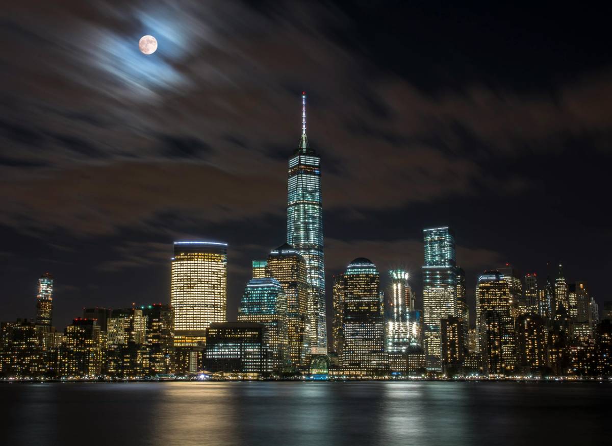 The Manhattan skyline at night viewed from across the New York Harbor, with illuminated skyscrapers and the moon shining through a partially cloudy sky.