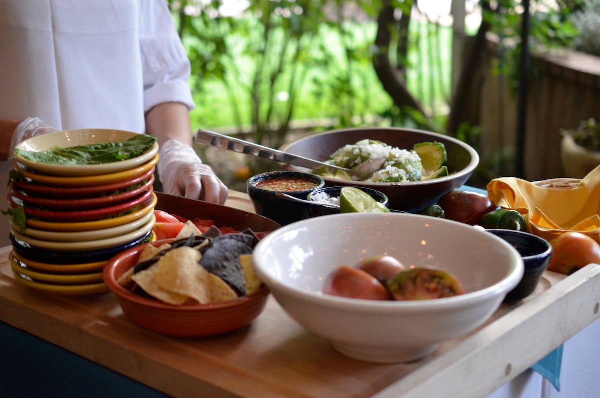 Colorful dishes filled with fresh ingredients, tortilla chips, salsa, and other Mexican food items on a wooden table, with a person preparing the meal in the background. A culinary delight on a road trip to Santa Fe from Denver, Colorado.
