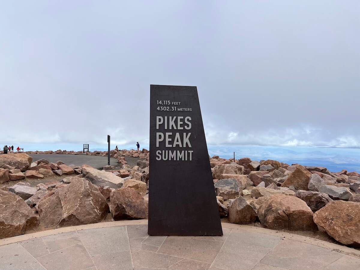 Sign at the Pikes Peak Summit reading '14,115 feet, 4302.31 meters, Pikes Peak Summit' surrounded by rocks with a cloudy sky in the background. Ideal for a road trip from Denver, Colorado.