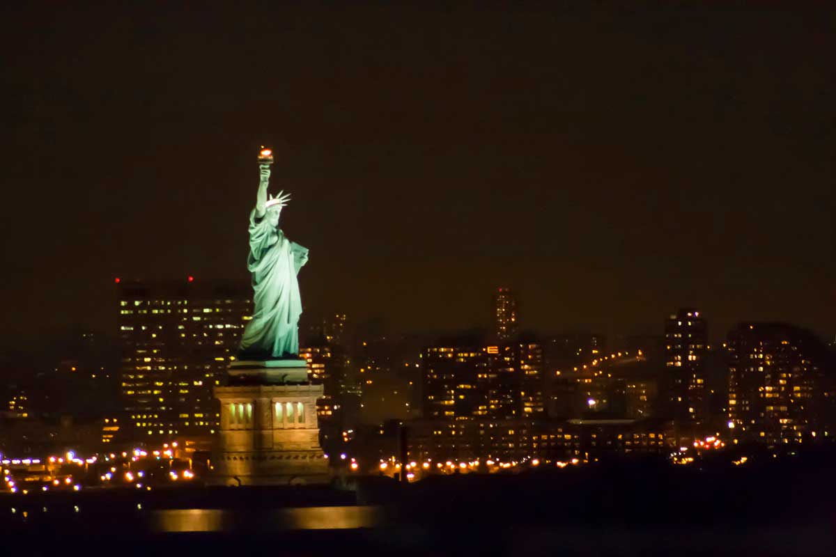 The Statue of Liberty at night, illuminated against the dark sky, as seen from the Staten Island Ferry with a few city lights visible in the background.