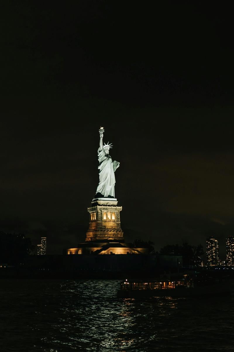 The Statue of Liberty at night, illuminated against the dark sky, as seen from the Staten Island Ferry with a few city lights visible in the background.