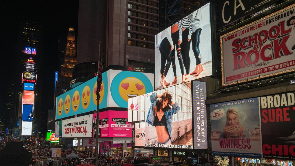 Times Square in Manhattan at night, showcasing vibrant billboards and advertisements, including images of emojis and fashion models, with crowds of people below.