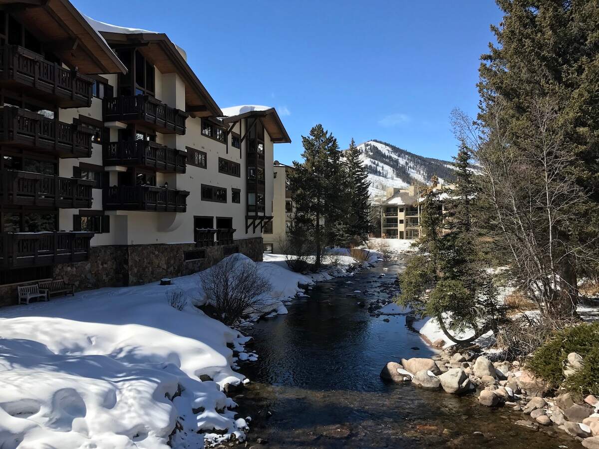 Charming buildings with wooden balconies in Vail Village, alongside a snow-covered landscape with a serene creek and snow-capped mountains in the background. A picturesque road trip destination from Denver, Colorado.