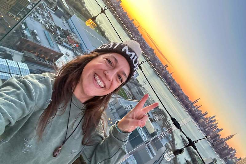 A woman smiling and making a peace sign at the Westlight Rooftop Bar in Brooklyn, with a stunning view of the Manhattan skyline at sunset just before nightfall.