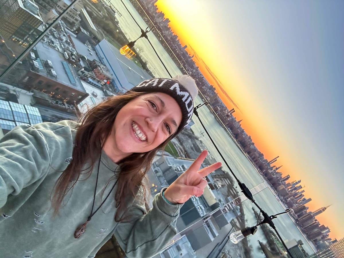 A woman smiling and making a peace sign at the Westlight Rooftop Bar in Brooklyn, with a stunning view of the Manhattan skyline at sunset just before nightfall.