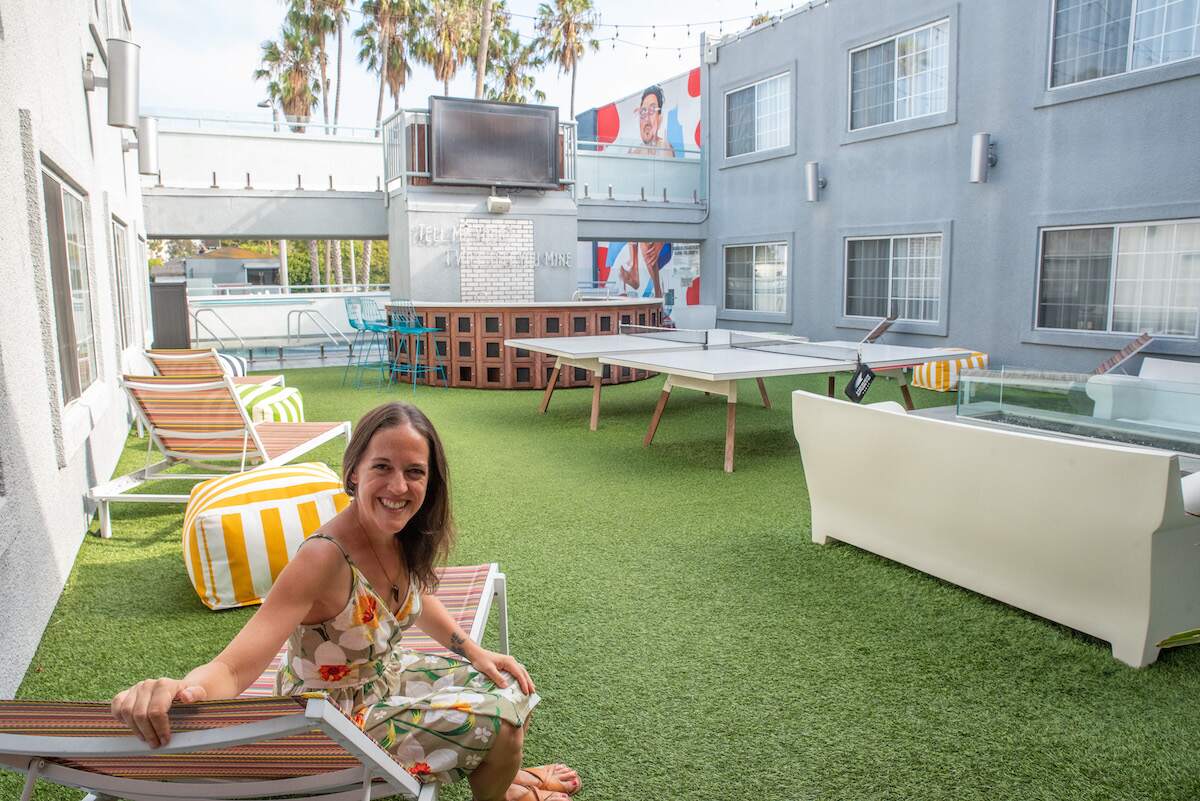 A female traveler in a green sundress sits comfortably on the eclectic rooftop of The Kinney Hotel in Venice, Los Angeles, California. Surrounding her are vibrant sun chairs, a sparkling pool, a bubbling hot tub, and colorful murals adorning the walls, creating a lively and inviting atmosphere.