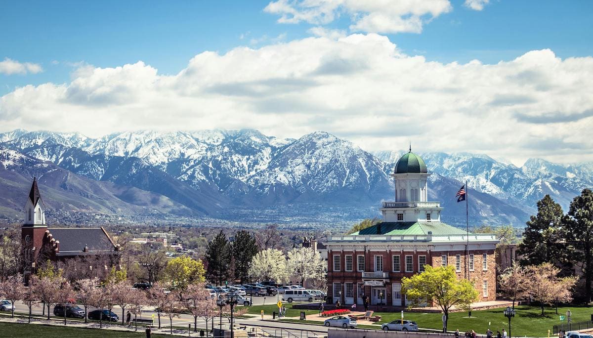 View of Salt Lake City with historic buildings and trees in the foreground, and the snow-capped Wasatch Mountains under a partly cloudy sky in the background. A picturesque road trip destination from Denver, Colorado.