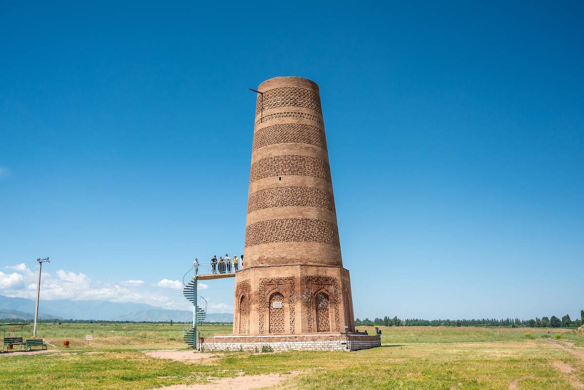 A tall, cylindrical brick tower known as Burana Tower stands prominently against a clear blue sky, with a spiral staircase leading up to a viewing platform where several people are standing. The tower, an ancient minaret, is surrounded by a vast, flat grassy landscape with distant mountains visible on the horizon. This historic site highlights the architectural heritage and scenic beauty of Kyrgyzstan.