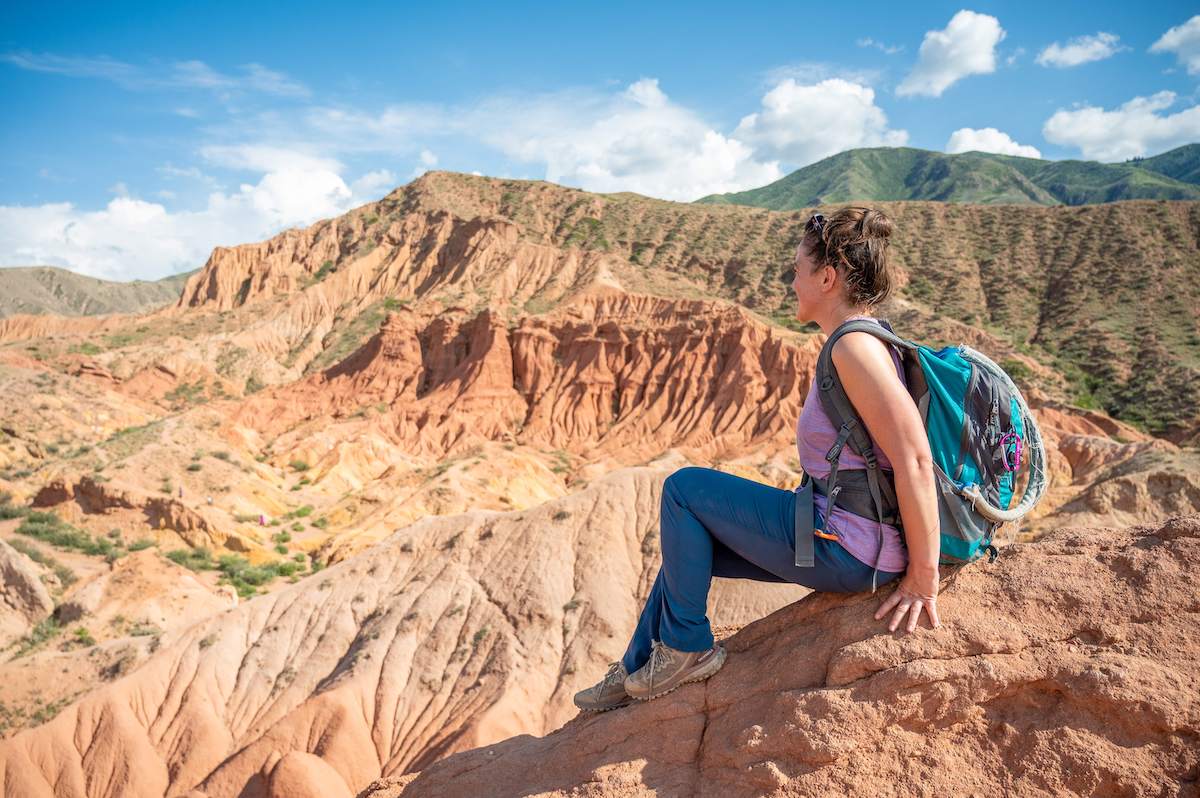 A woman sits on a rocky ledge in Fairytale Canyon, Kyrgyzstan, gazing out over the vibrant, eroded sandstone cliffs and ridges that stretch into the distance under a bright blue sky. She is wearing a backpack and casual hiking attire, capturing a moment of peaceful reflection amidst the canyon's stunning and colorful landscape.