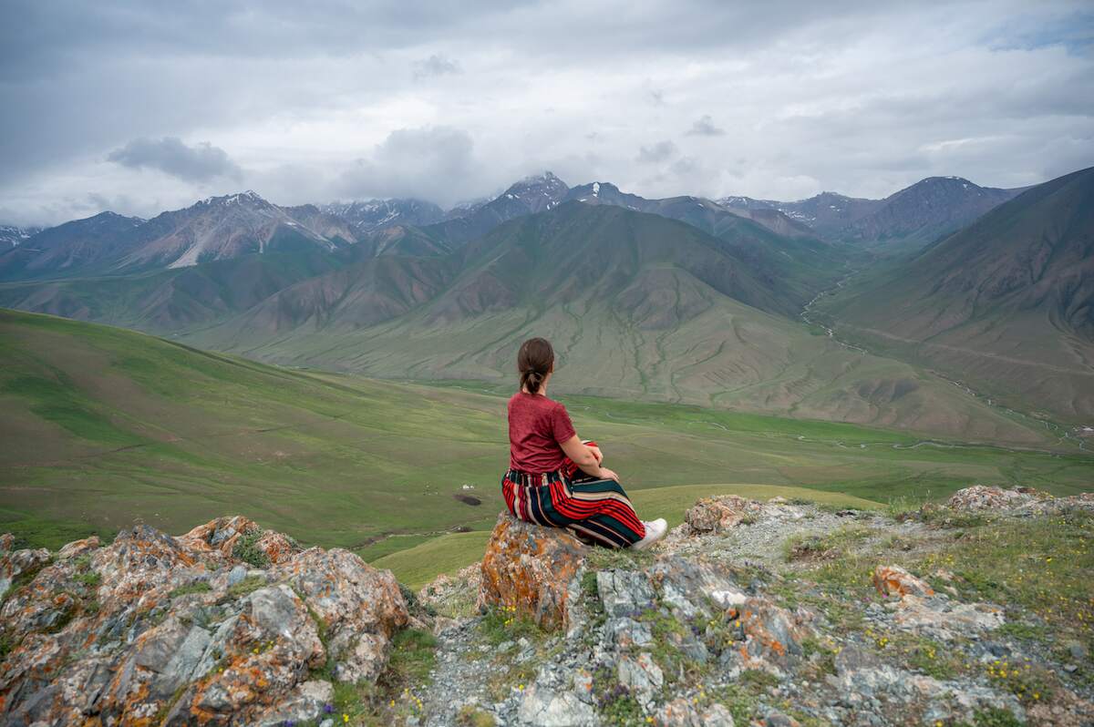 A traveler sits on a rock, gazing at the expansive views from Kalmak Ashuu Pass, where rolling green hills meet rugged mountain peaks under a cloudy sky, capturing the essence of a 14-day adventure in Kyrgyzstan.