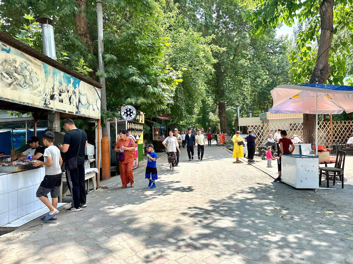 A vibrant scene at Alisher Navoi Park in Osh City, Kyrgyzstan, featuring a tree-lined pathway with various food stalls and people leisurely walking, eating, and socializing on a sunny day. The park is bustling with activity, showcasing local vendors serving traditional snacks and families enjoying the pleasant outdoor environment.