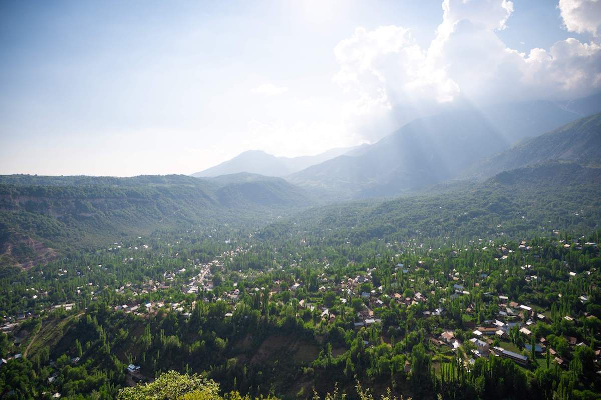 Panoramic viewpoint over Arslanbob Village showcasing a lush green valley dotted with houses and trees, with mountains and sunlight streaming through clouds in the background.