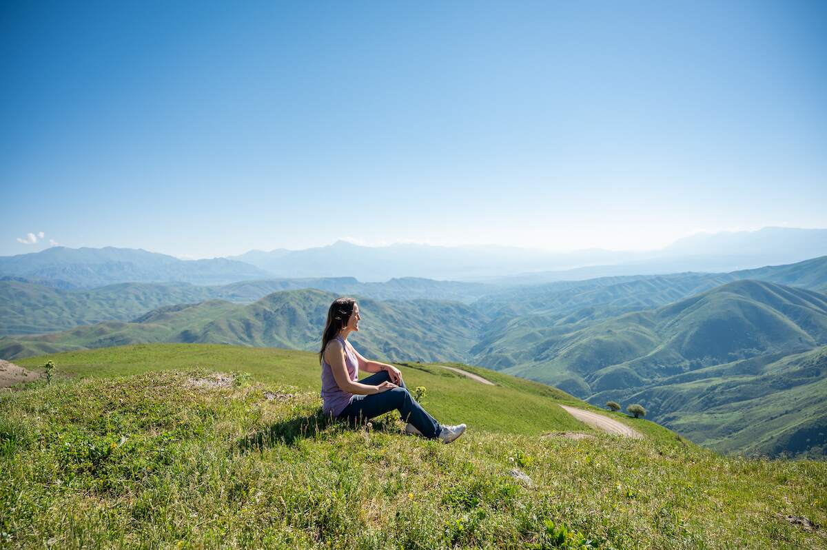 A serene view from the Asylbek Tologonov Tuz Ashuu Pass, showcasing a traveler sitting peacefully on the lush green hillside, with rolling mountains and a clear blue sky in the background, capturing the essence of traveling to Kyrgyzstan.