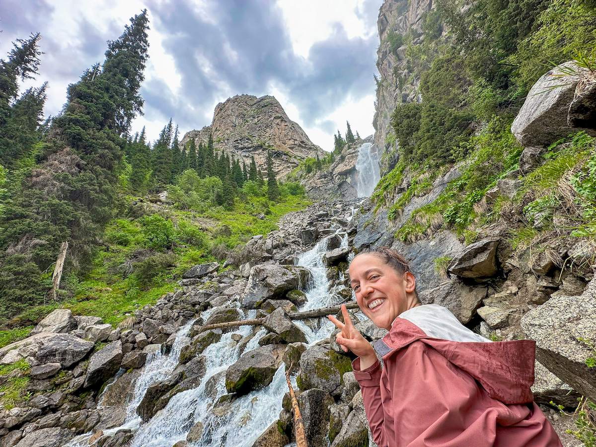 A joyful hiker poses with a peace sign in front of the cascading Barskoon Waterfall in Kyrgyzstan, surrounded by lush greenery, rocky terrain, and towering pine trees under a dramatic cloudy sky. This scenic spot is a highlight for adventurers and nature lovers exploring the country's natural wonders during their Kyrgyzstan travel itinerary.