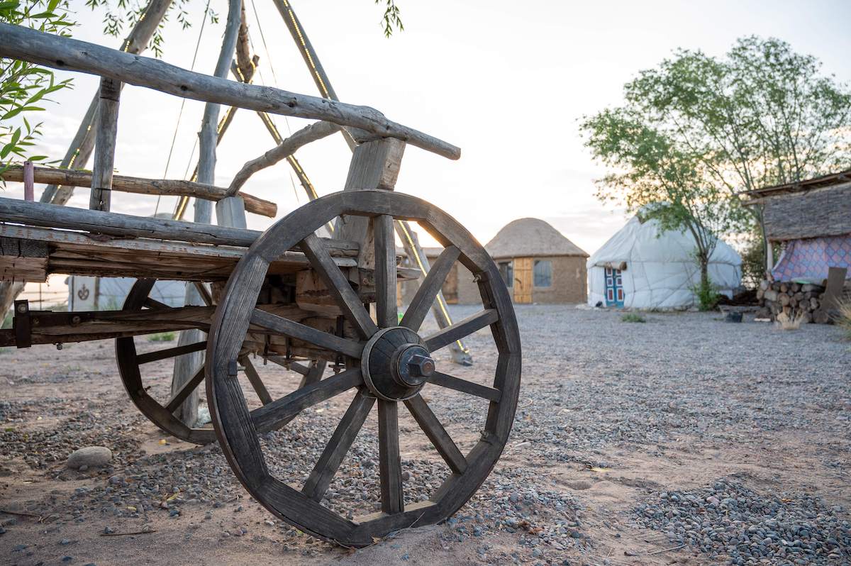 A rustic wooden cart wheel stands in the foreground at Bell Tam Yurt Camp on Issyk Kul Lake, Kyrgyzstan, with traditional yurts and a cozy mud hut visible in the background against the soft light of sunset. This camp offers an authentic and serene experience for those traveling in Kyrgyzstan.
