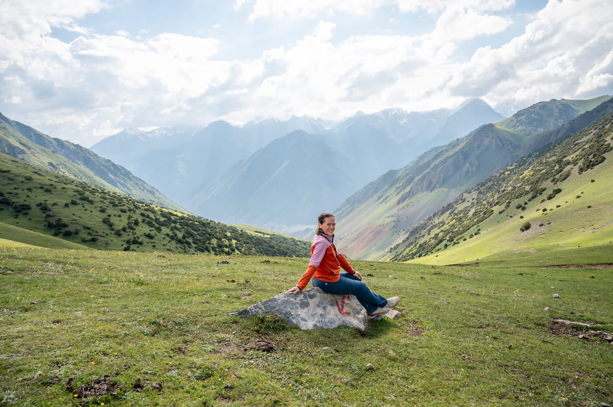 A woman sitting on a rock at Kosh Moinok Pass, enjoying the stunning view of green valleys and distant mountain peaks under a partly cloudy sky. The scenery highlights the natural beauty of Kyrgyzstan.