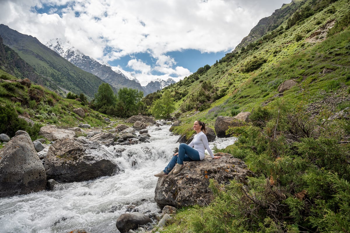 A woman sitting on a rock beside a rushing mountain stream during a hike to Kosh Moinok Pass in Kyrgyzstan, surrounded by lush greenery and snow-capped peaks in the background.