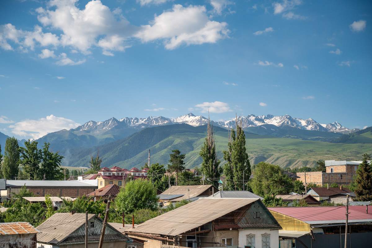 A breathtaking view of snow-capped mountains from the rooftop of the Mongu Hotel in Karakol, Kyrgyzstan, with the town's rooftops and lush greenery in the foreground. This picturesque scene captures the stunning natural beauty and serene atmosphere travelers can enjoy while planning a trip to Kyrgyzstan.