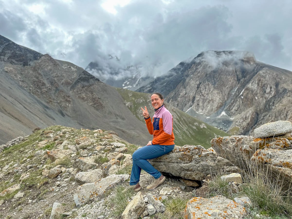 A hiker wearing a colorful jacket and blue pants sits on a rock, flashing a peace sign, while enjoying the dramatic mountain views along the Sary Mogul trek in Kyrgyzstan.
