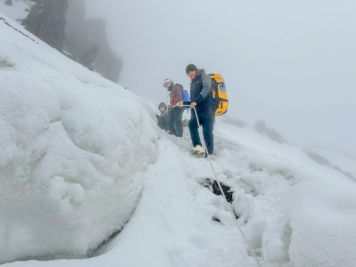 A group of climbers carefully navigates a steep, snowy slope down from  Sary Mogul Pass in Kyrgyzstan, roped together for safety and carrying heavy backpacks amidst foggy, challenging conditions. This scene captures the adventurous spirit and physical demands of trekking in Kyrgyzstan, ideal for those seeking high-altitude experiences during their travel itinerary.