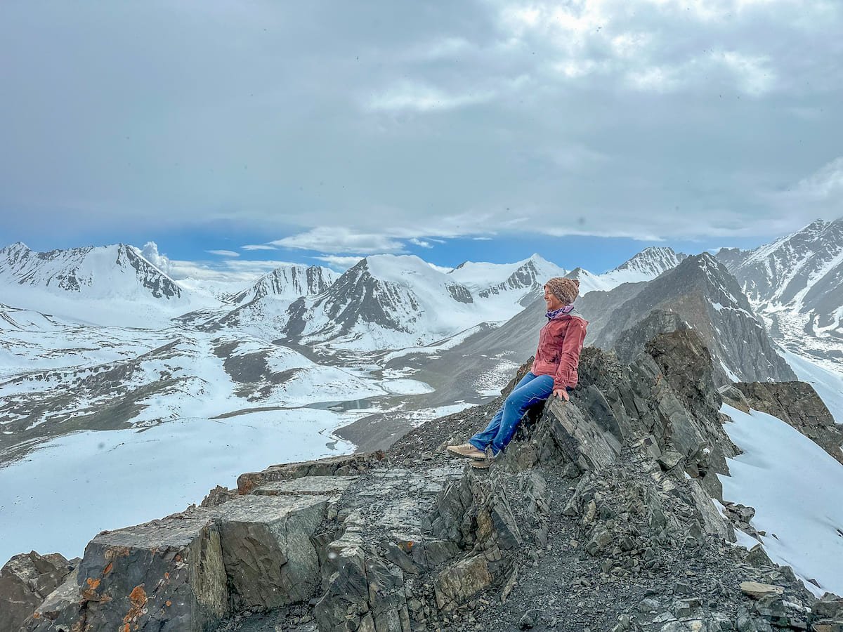 A hiker sits on a rocky outcrop, admiring the snow-covered peaks and expansive snowy landscape from Mary Mogul Pass in Kyrgyzstan.