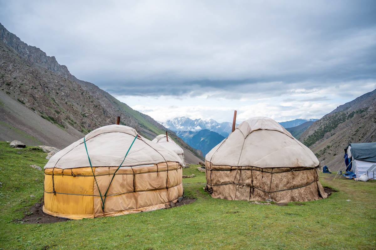 Two traditional yurts at Sary Mogul Yurt Camp in Kyrgyzstan, surrounded by green pastures and rugged mountain scenery under a cloudy sky.