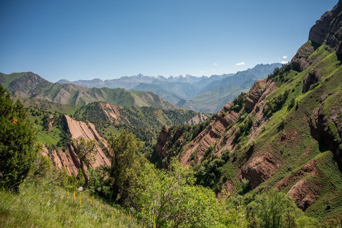 A view of the rugged landscape at Three Dragon Gorge, featuring steep, rocky cliffs and verdant greenery. The background reveals distant mountain ranges under a clear blue sky, showcasing the natural beauty of the Kyrgyzstan region.