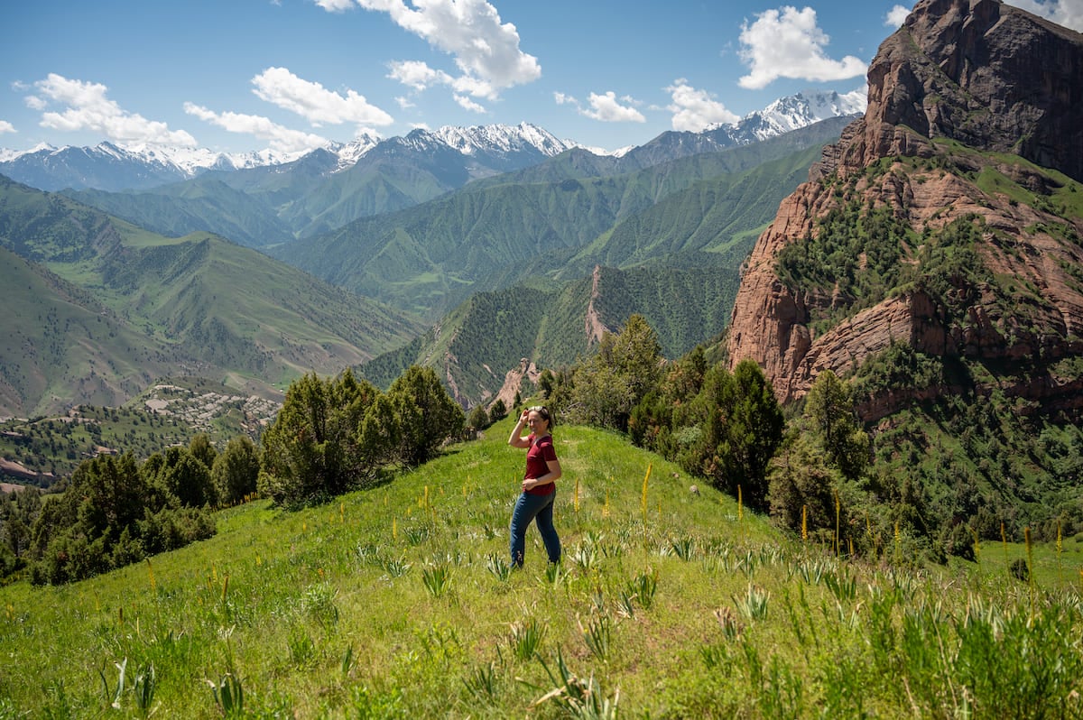 A woman standing on a grassy hillside, surrounded by lush green vegetation, with majestic snow-capped mountains and rocky formations in the background, taken while trekking in Kyrgyzstan.