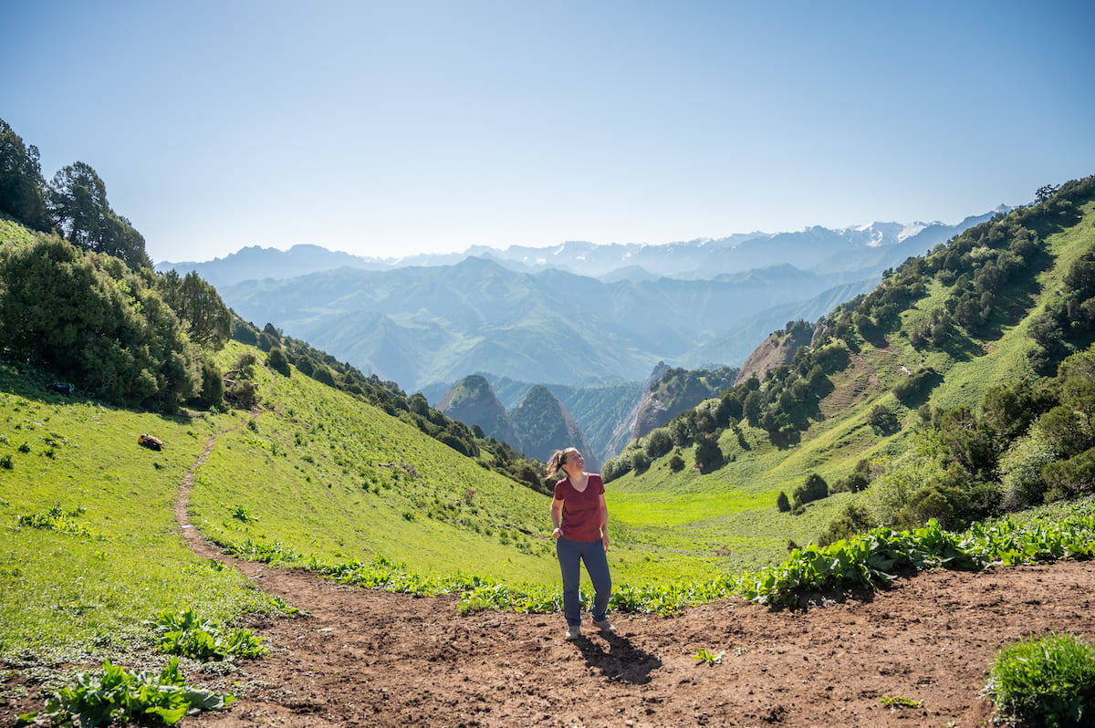 A woman standing on a dirt path at Sarybell Pass, surrounded by lush green hills and valleys. The distant mountain ranges stretch under a clear blue sky, showcasing the breathtaking landscape of Kyrgyzstan.