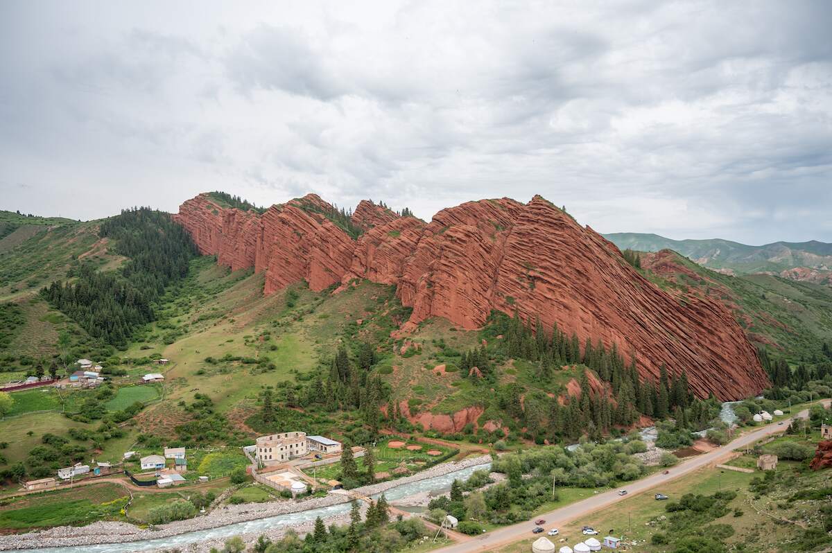 The striking formation of 7 Bulls Rock in Kyrgyzstan, showcasing vibrant red sandstone cliffs that resemble a line of bulls, set against a backdrop of green hills and a cloudy sky. Below the formation, a small village with houses and a river add to the picturesque landscape, making this a must-see spot for travelers exploring Kyrgyzstan.