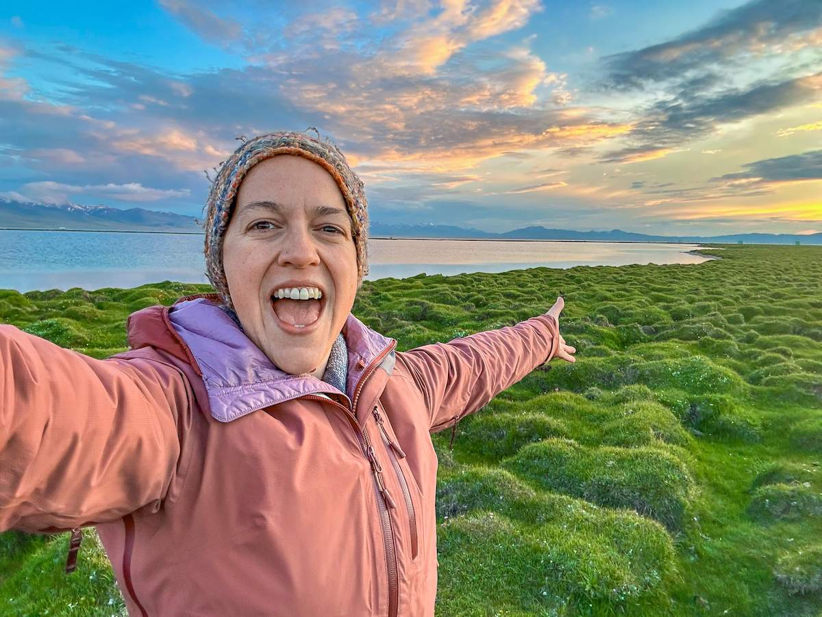 A traveler joyfully poses with outstretched arms, capturing the breathtaking sunset over Song Kul Lake, where the sky's vibrant hues reflect on the serene water, and lush green grass stretches along the shoreline, highlighting the beauty of a Kyrgyzstan travel itinerary.
