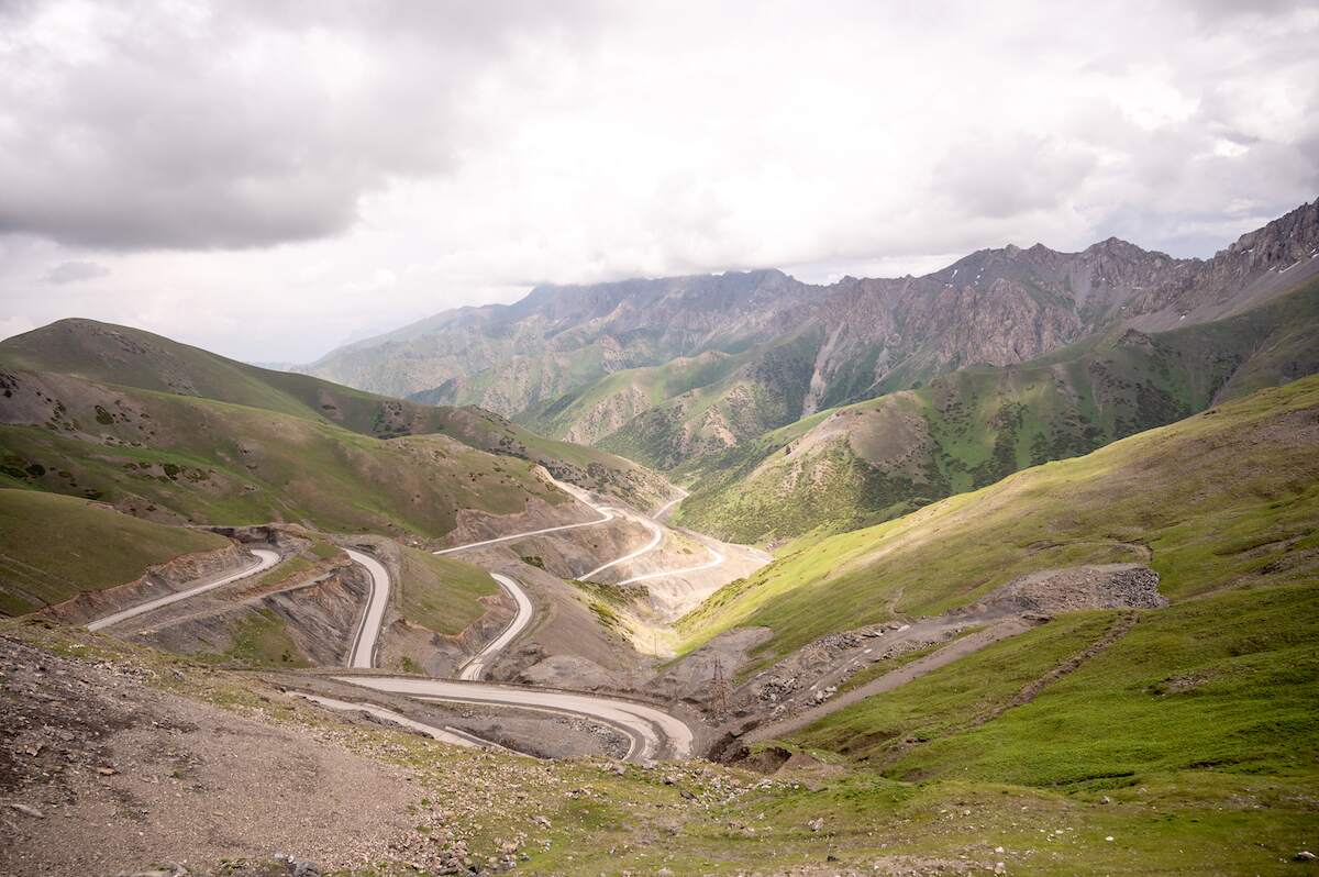 A panoramic view from Taldyk Pass along the Pamir Highway, showcasing winding roads cutting through the rugged, green mountain landscape under a cloudy sky.