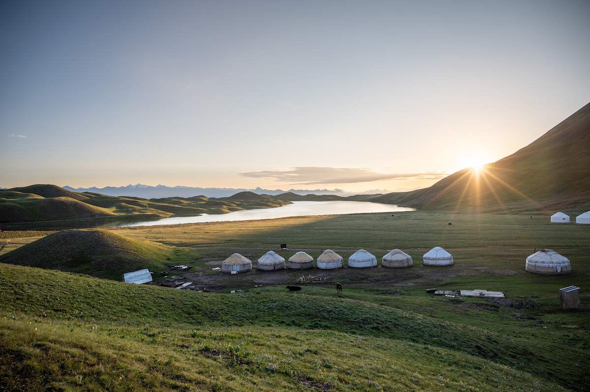 The view over Tulpar Kul Lake at sunrise, featuring a row of yurts in the foreground, gentle hills, and the sun rising behind a mountain, casting a golden glow over the serene landscape.