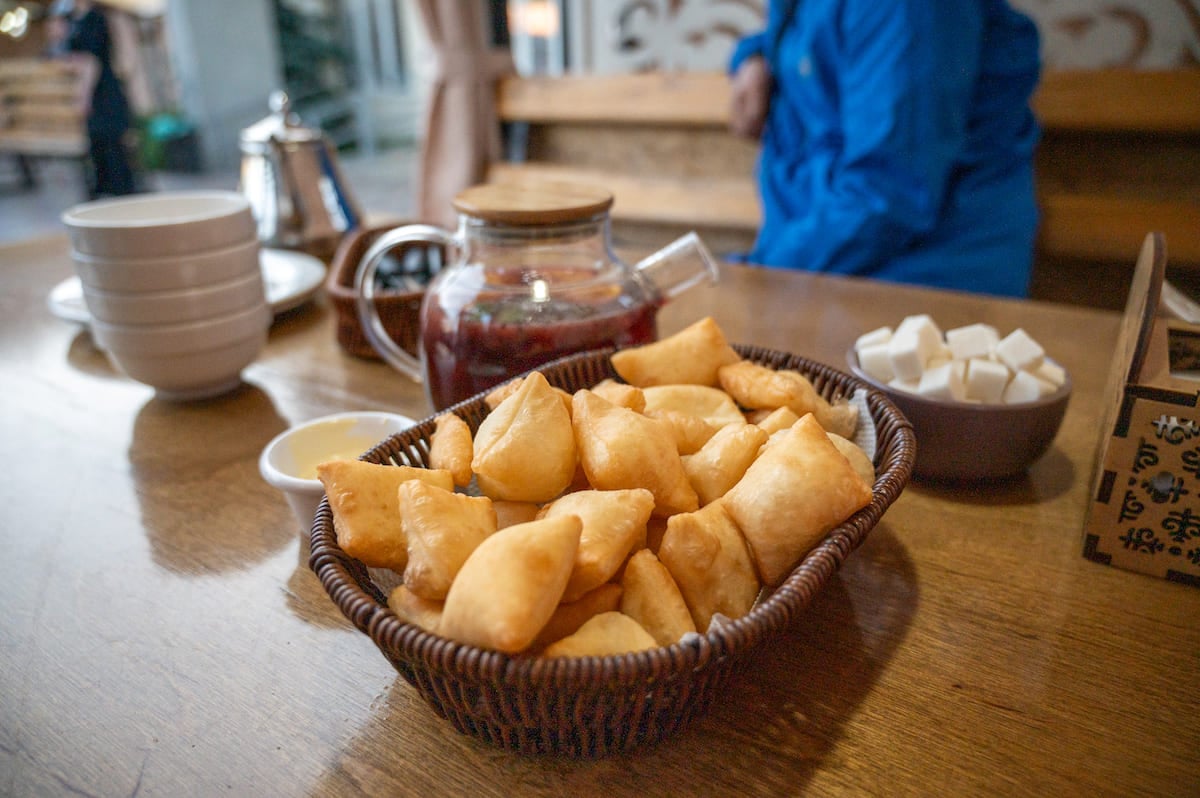 A close-up shot of a wicker basket filled with freshly fried borsook, which are golden-brown dough pieces. Beside the basket, there is a glass teapot filled with dark hot tea, a bowl of white sugar cubes, and a few white bowls stacked together, all placed on a wooden table, suggesting a cozy and inviting setting.