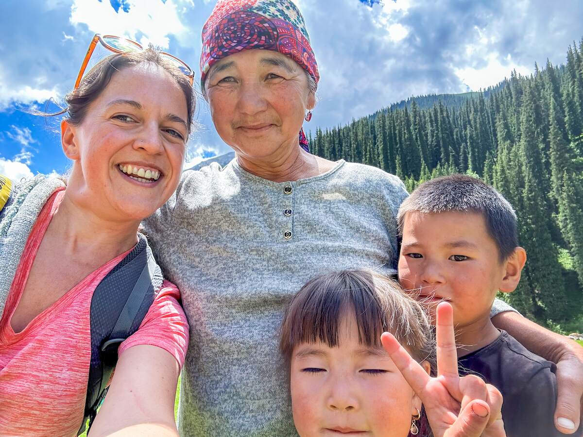 A joyful selfie with a local Kyrgyz family who offered tea and snacks to hikers, featuring a smiling woman in a bright pink shirt alongside an older woman wearing a colorful headscarf and two young children. The group stands against a backdrop of lush green pine trees and a clear blue sky, capturing a moment of warm hospitality and cultural exchange during the hiking adventure.