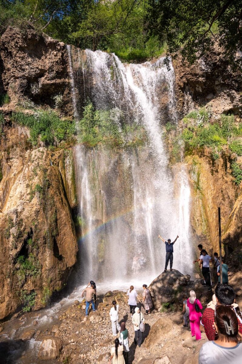 A group of people enjoying the refreshing mist and the scenic beauty of the Small Arslanbob Waterfall in Arslanbob Village, with a vibrant rainbow visible through the cascading water.