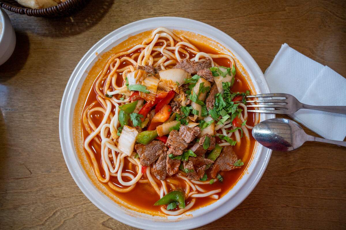 A vibrant bowl of Laghman, a popular dish in Kyrgyzstan, featuring hand-pulled noodles in a rich, red broth, topped with tender slices of beef, bell peppers, onions, and fresh herbs. The colorful presentation and aromatic ingredients highlight the hearty and flavorful nature of this traditional Central Asian cuisine. A fork and spoon rest beside the bowl on a wooden table, inviting the viewer to enjoy this delicious meal.