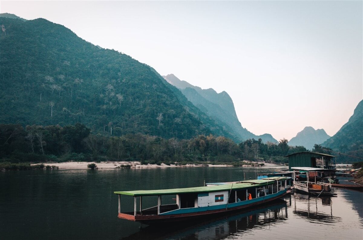 Traditional local boats docked along a serene river in Laos, with lush green mountains forming a stunning backdrop, illustrating the charm of solo travel in Laos.