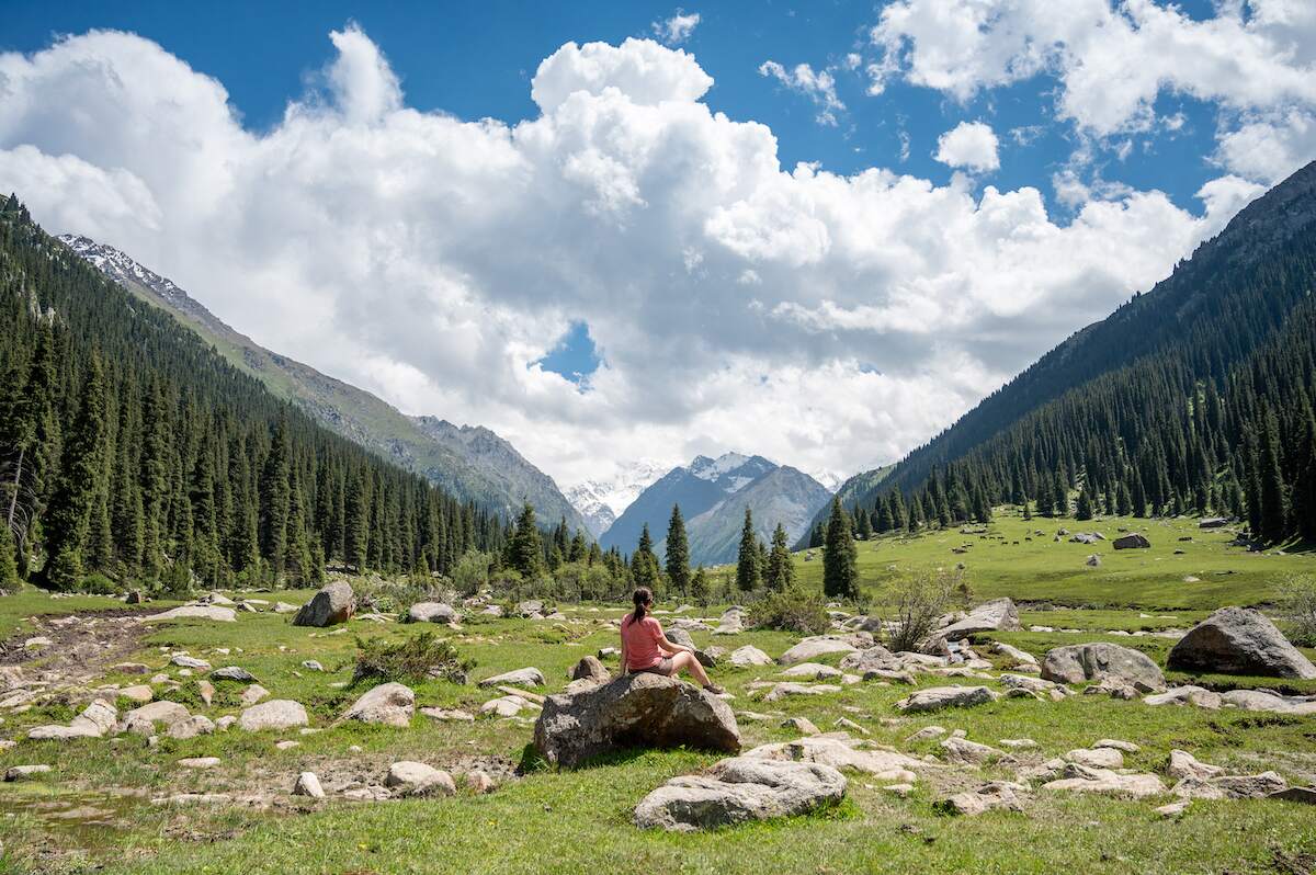 A scenic view of Oguz-Bashi Peak (Yeltsin Peak) in the Tian Shan Mountain Range, featuring a lush, green valley filled with scattered rocks and surrounded by dense pine forests. In the foreground, a woman sits on a large rock, admiring the snow-capped peak and the dramatic, cloud-filled sky above. The tranquil and picturesque setting captures the natural beauty and serene atmosphere of the Kyrgyzstan landscape.