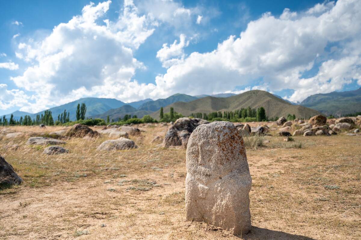 An ancient Turkic stone sculpture stands prominently in the Open Air Petroglyph Museum in Cholpon-Ata, Kyrgyzstan, amidst a field scattered with other stones and petroglyphs. The backdrop features rolling hills, tall trees, and a partly cloudy sky, capturing the historical and cultural significance of this site, a highlight in any Kyrgyzstan travel guide.