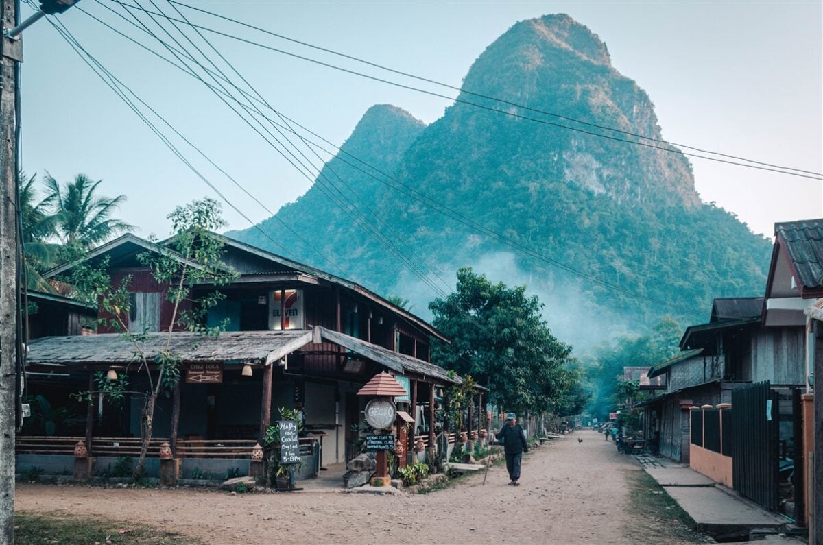 Solo traveler in Laos walking down a quaint street in Muang Ngoi lined with traditional wooden houses and surrounded by towering green mountains.